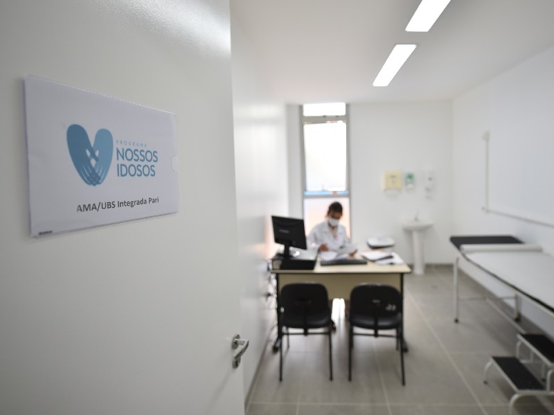 Consultório médico com paredes brancas. A porta está entreaberta e ao fundo se vê uma mulher trabalhando numa mesa com computador. Do lado direito há uma maca e ao fundo uma pia